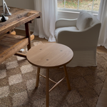 A round wooden side table with three legs and a natural light wood finish, positioned in a cozy room. Traditional joinery white oak cricket table.