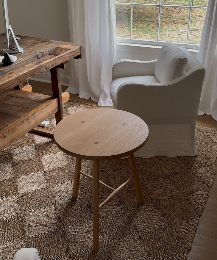 A round wooden side table with three legs and a natural light wood finish, positioned in a cozy room. Traditional joinery white oak cricket table.