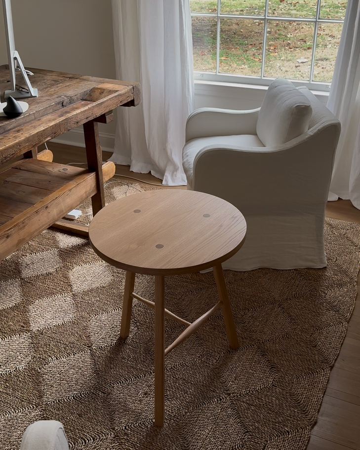 A round wooden side table with three legs and a natural light wood finish, positioned in a cozy room. Traditional joinery white oak cricket table.